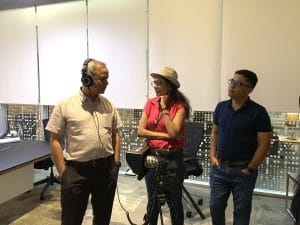 Group discussing in office with camera equipment; one person wearing headphones, others in casual attire.