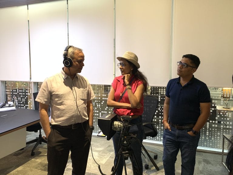 Group discussing in office with camera equipment; one person wearing headphones, others in casual attire.